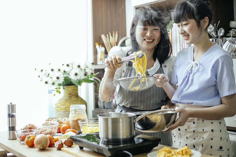 Cheerful Asian teen girl with middle aged mother draining boiled pasta in stainless bowl cooking in team